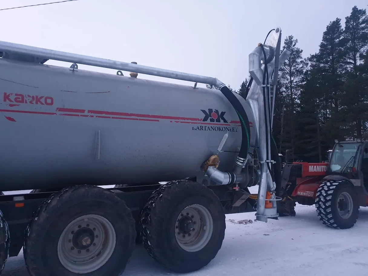 Suurikokoinen peltisäiliö, jossa on mustia letkuja ja renkaat lumisella pihalla, taustalla traktori ja mäntyjä.