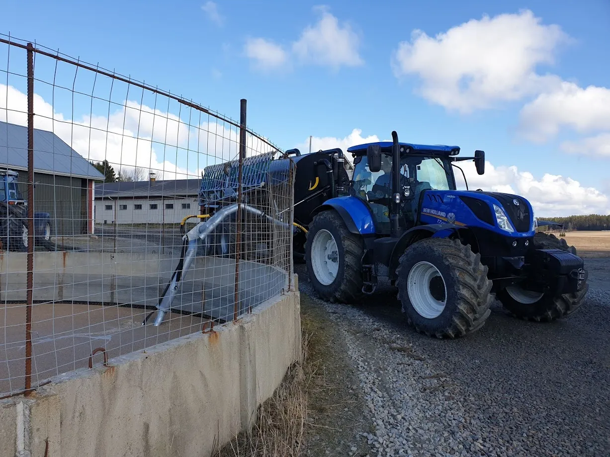 Sininen New Holland -traktori maatalousalueella, joka on kytketty rehuvaunuun aitan lähellä.