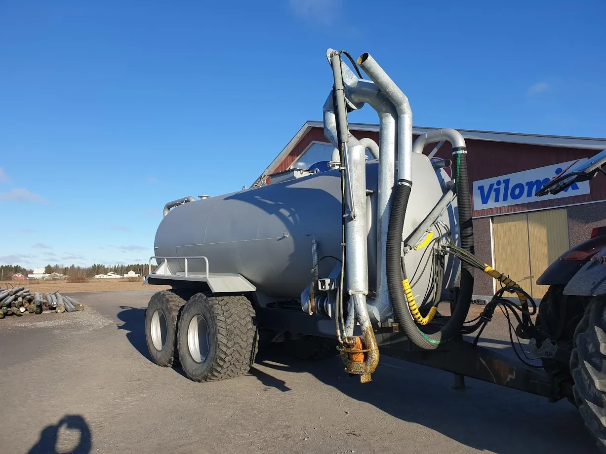 Metallinen lietevaunu, jossa on suuret renkaat ja putkisto, yhdistettynä traktoriin ulkona aukiolla sinisen taivaan alla.