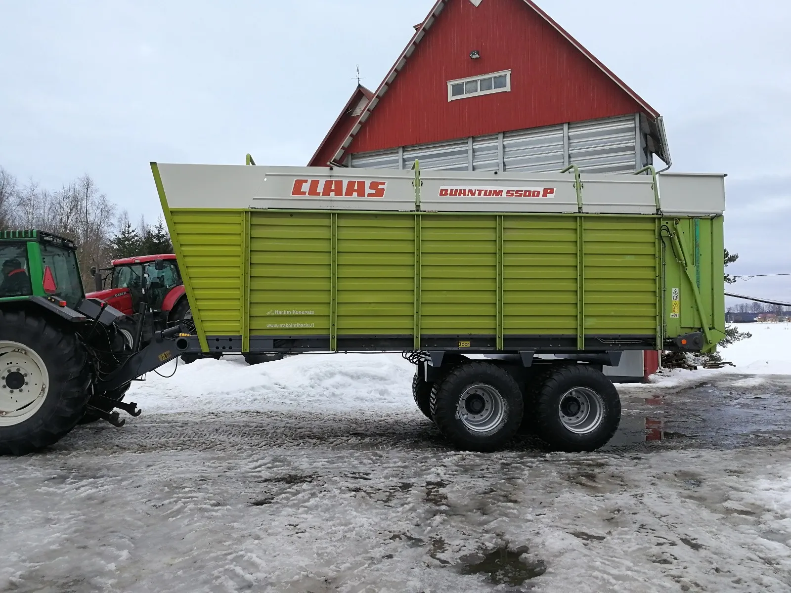 Vihreä CLAAS Quantum 5500 P -rehukärry kytketty traktoriin lumisella pihamaalla punaisen rakennuksen edessä.