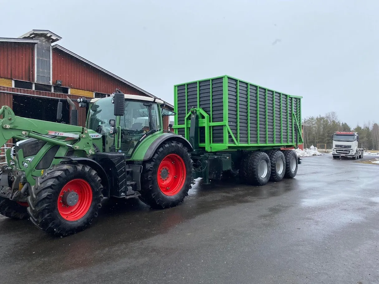 Vihreä traktori punaisilla pyörillä ja perässä vihreä maatalouskärry, taustalla punainen lato ja valkoinen kuorma-auto.
