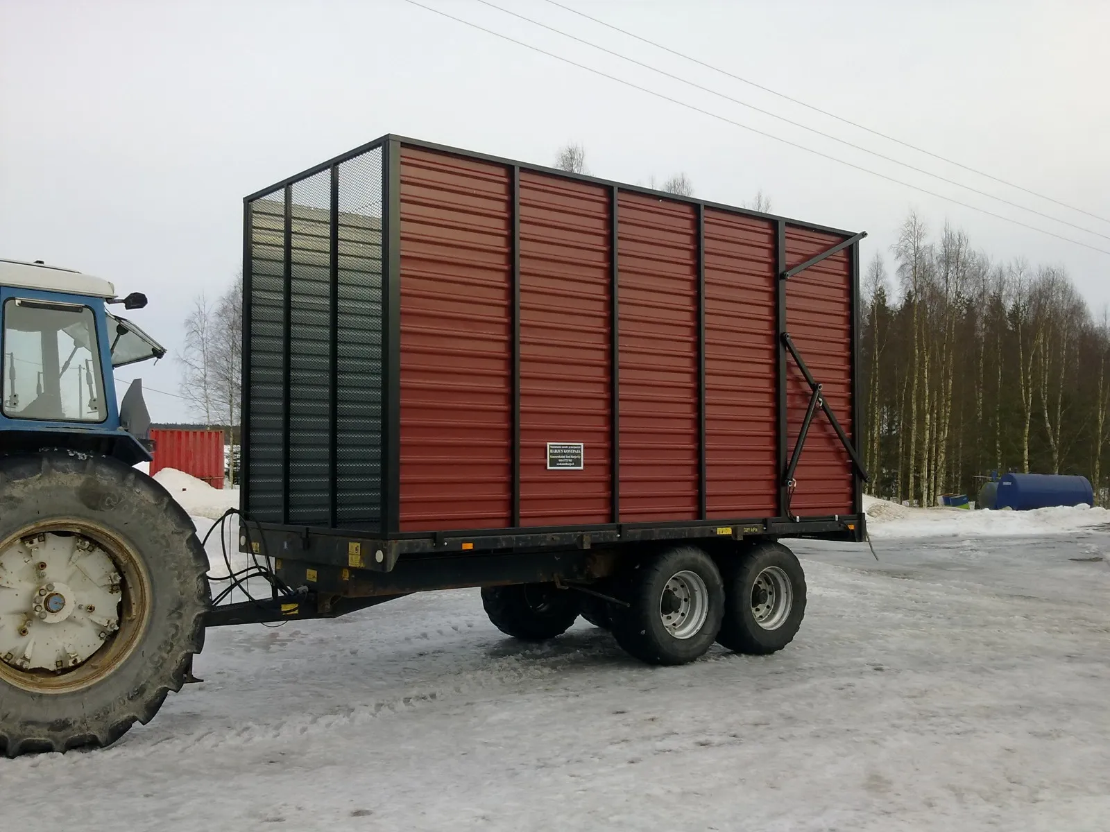 Sininen traktori vetää suurta kahdella akselilla varustettua peräkärryä, jossa on punaiset sivuseinät ja musta kehys lumisella pihamaalla.
