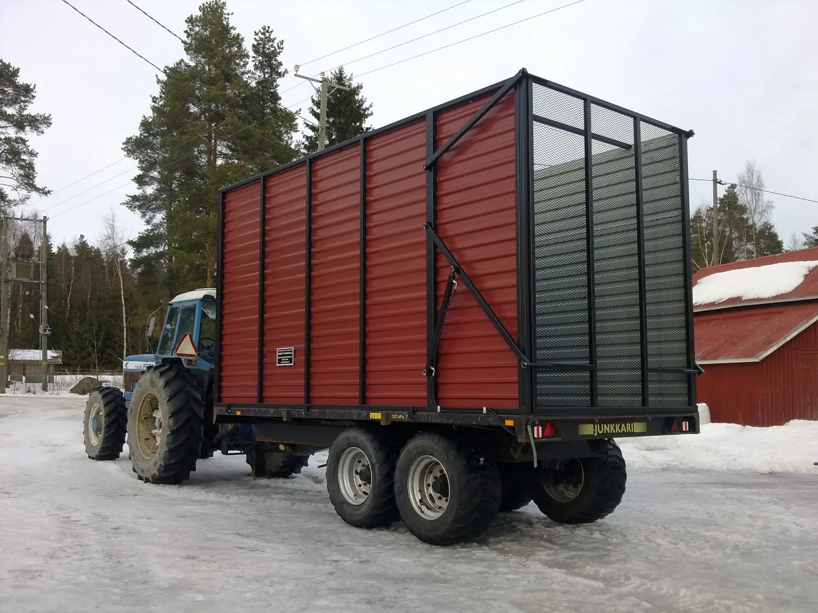 Sinivalkoinen traktori vetää punaista Junkkari-merkkistä peräkärryä lumisella pihalla.