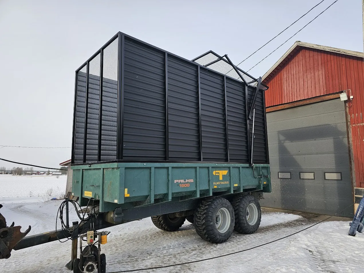 Metallinen maansiirto- ja kuljetusvaunu lumisella pihalla punaisen rakennuksen vieressä.