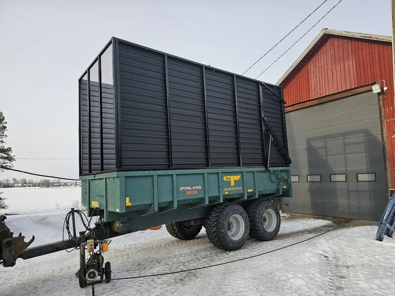 Vihreä maatalouskärry mustalla korilla lumisella pihalla punaisen rakennuksen vieressä.