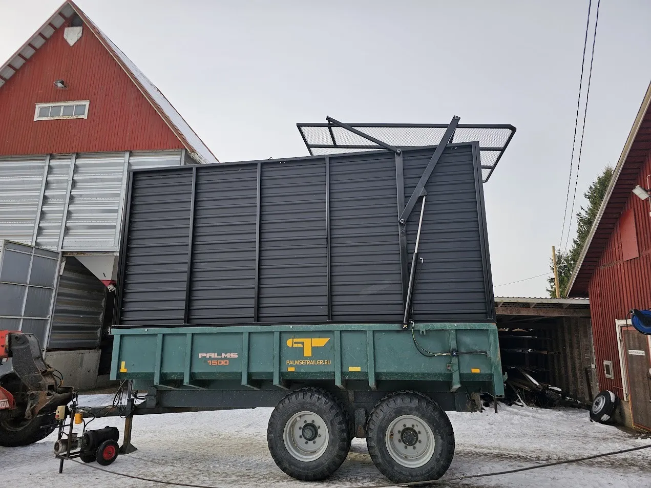 Vihreä ja musta Palms 1500 maansiirtovaunu lumisella pihalla maatalousrakennusten välissä.
