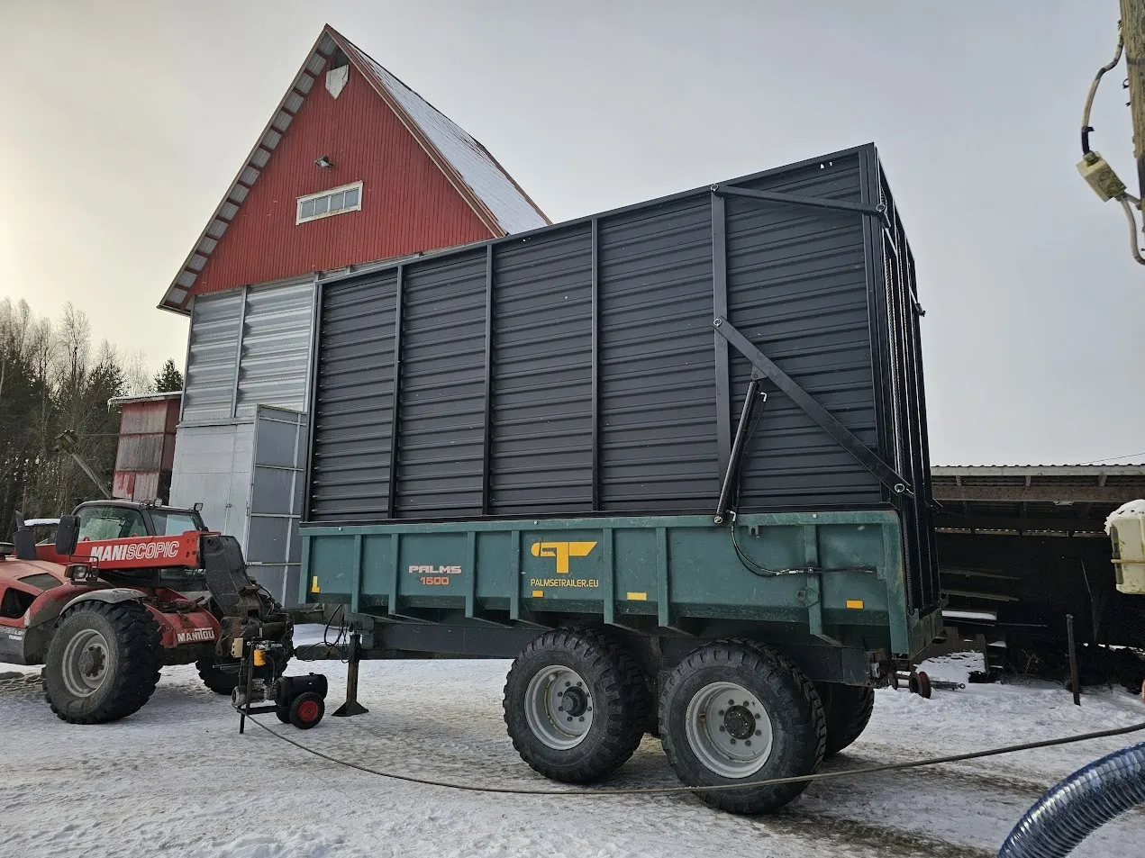 Traktori vetää Palms 1500 -peräkärryä pihalla lumisessa miljöössä, taustalla punainen lato.
