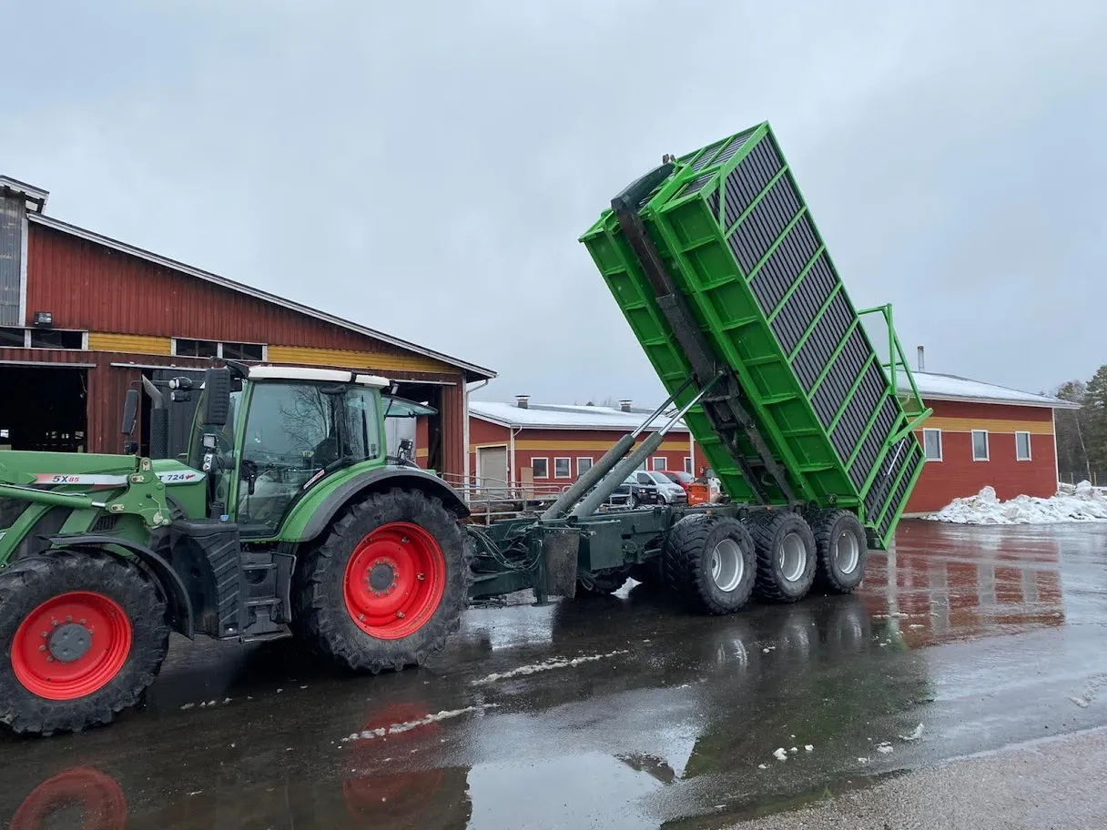 Vihreä traktori, jossa on suuri vihreä perävaunu nostettuna, pihalla märällä asfalttipinnalla maatilan rakennusten edessä.