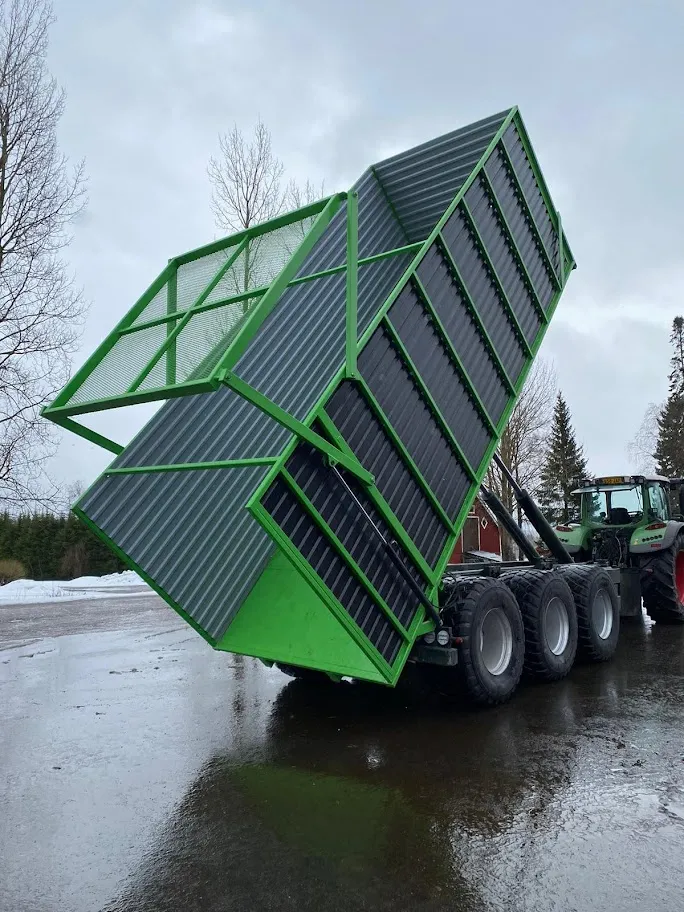 Vihreä traktorin perävaunu on nostettu ylös, taustalla lehtiä pudottavat puut ja märkä asfaltti.