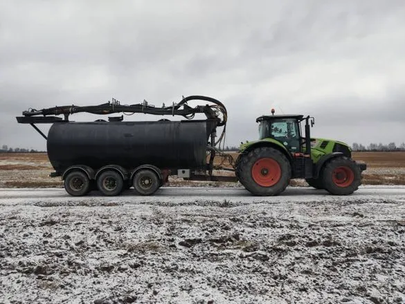 Vihreä traktori, jossa punaiset pyörät, vetää mustaa kolmirenkaista säiliövaunua lumisella maatalouspellolla.